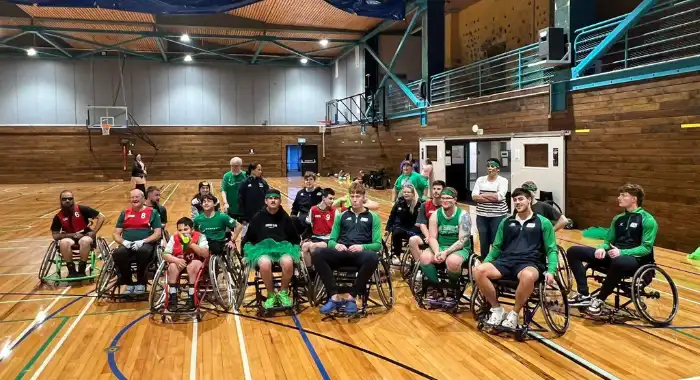 A group of people, including kids and adults, are gathered in a gym, many in wheelchairs, wearing sports jerseys. They’re seated in a semi-circle on a basketball court, smiling and enjoying their time together after what seems to be a fun sports event. The setting is warm and inclusive, with a basketball hoop in the background.